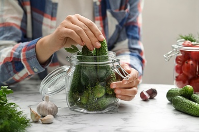 Woman putting cucumber into glass jar at white marble kitchen table, closeup. Pickling vegetables