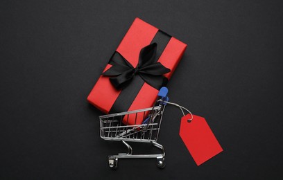 Shopping cart with price tag and gift box on black background, flat lay