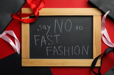 Small chalkboard with phrase SAY NO TO FAST FASHION and black shopping bags on red background, flat lay