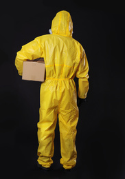 Man wearing chemical protective suit with cardboard box on black background, back view. Prevention of virus spread