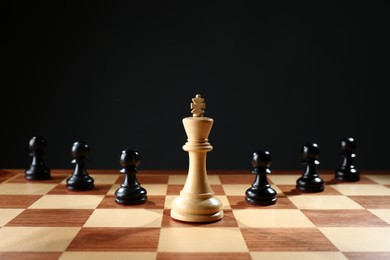 White king among black pawns on wooden chess board against dark background