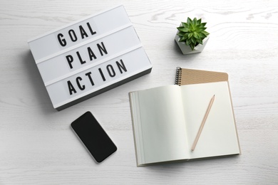 Light box with text Goal, Plan, Action near notebooks and smartphone on white wooden table, flat lay
