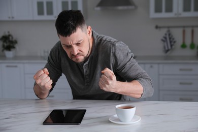 Emotional man with tablet at table in kitchen. Online hate concept