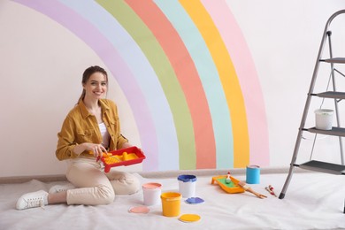 Photo of Young woman holding tray and roller near wall with painted rainbow indoors