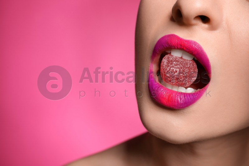 Young woman with beautiful makeup holding candy in lips on pink background, closeup. Space for text