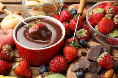Fondue fork with strawberry in bowl of melted chocolate surrounded by other fruits on wooden table