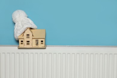 Modern radiator with knitted hat and wooden house near light blue wall indoors, space for text. Winter heating efficiency
