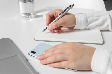 Woman with smartphone writing in notebook at white table, closeup