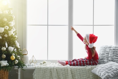 Photo of Cute little girl in Santa hat on window sill near Christmas tree at home