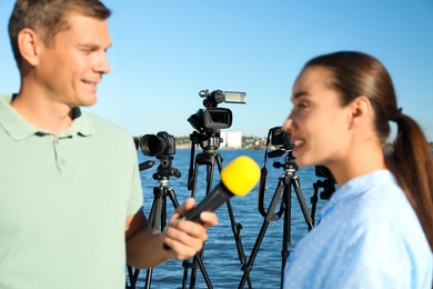 Journalist interviewing young woman near river, focus on video cameras