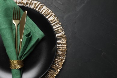 Photo of Plate with green fabric napkin, decorative ring and cutlery on black table, top view. Space for text