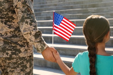 Soldier and his little daughter with American flag holding hands outdoors, back view. Veterans Day in USA
