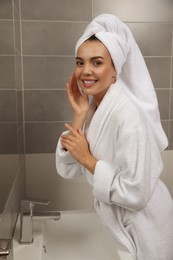 Beautiful young woman in bathrobe with towel on head near mirror at home