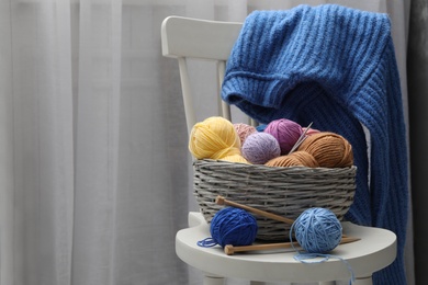 Photo of Woolen yarns in basket and knitting needles on chair indoors. Space for text