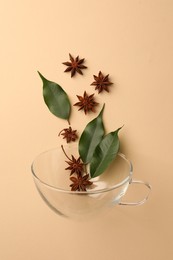 Photo of Anise stars and green leaves falling into glass cup on beige background, flat lay