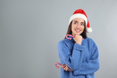 Young woman in blue sweater and Santa hat holding candy canes on grey background, space for text. Celebrating Christmas