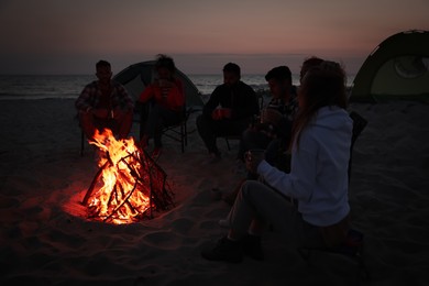 Group of friends gathering around bonfire on beach in evening. Camping season