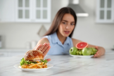 Woman choosing between fruits and burger with French fries in kitchen, focus on food