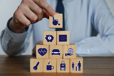 Man building pyramid of cubes with different icons at wooden table, closeup. Insurance concept