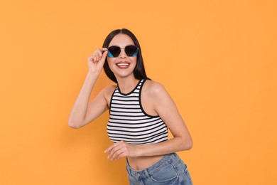 Attractive happy woman touching fashionable sunglasses against orange background