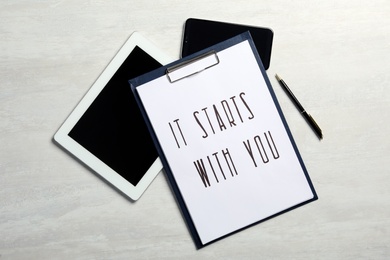 Clipboard with phrase It Starts With You, tablet and smartphone on white table , flat lay