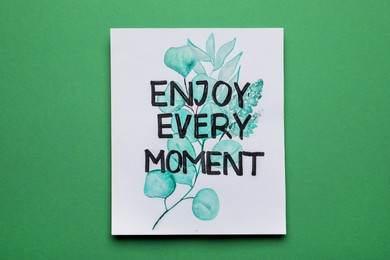 Card with beautiful phrase Enjoy Every Moment on green background, top view