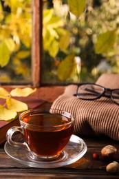 Photo of Cup of aromatic tea, acorns and soft sweater on wooden windowsill indoors. Autumn atmosphere