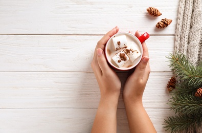Woman holding cup of tasty cocoa with marshmallows on white wooden table, top view. Space for text