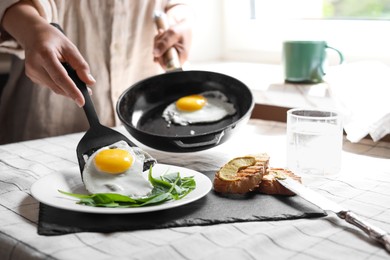 Woman putting tasty fried eggs onto plate at table indoors, closeup