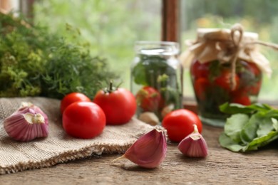 Photo of Glass jars, fresh vegetables and herbs on wooden table indoors, focus on garlic. Pickling recipe