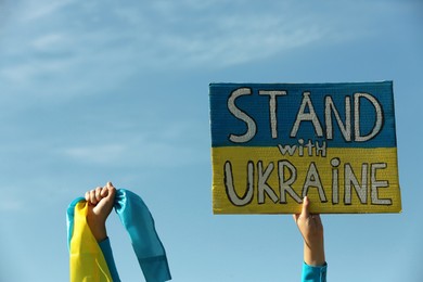 Woman holding poster Stand with Ukraine and national flag against blue sky, closeup