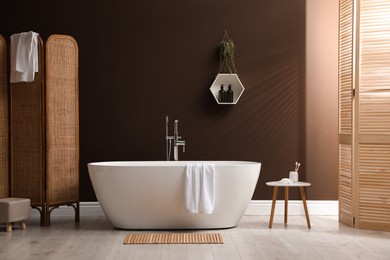 Modern ceramic bathtub with towel near brown wall in room