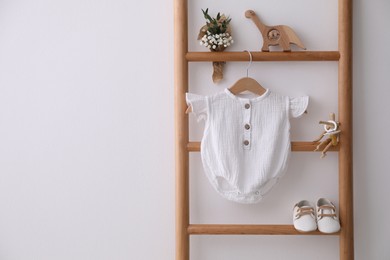Baby bodysuit, shoes, toys and small bouquet on ladder near white wall. Space for text