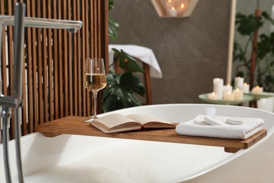 Wooden bath tray with glass of wine, open book, massage stones and towel on tub indoors. Relaxing atmosphere