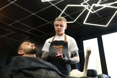 Professional hairdresser with shaving foam near bearded client in barbershop, low angle view