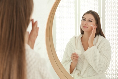 Young woman applying face cream onto her face near mirror in bathroom