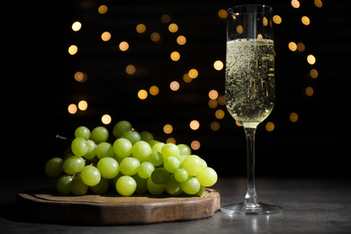 Green grape and glass of champagne on grey table against black background with blurred lights. Bokeh effect