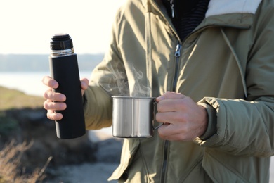 Photo of Man holding thermos and aluminium mug with hot drink outdoors, closeup