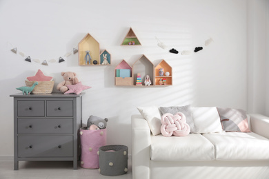 Photo of Cute children's room with house shaped shelves, sofa and chest of drawers. Interior design