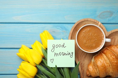 Cup of aromatic coffee with croissant, beautiful yellow tulips and Good Morning note on light blue wooden table, flat lay. Space for text