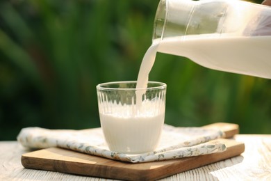 Pouring tasty fresh milk from bottle into glass on white wooden table, closeup