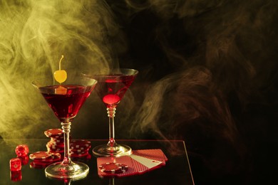 Casino chips, dice, playing cards and cocktails on dark background with smoke, space for text