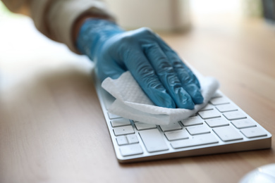 Woman in latex gloves cleaning computer keyboard with wet wipe at table, closeup