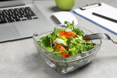 Photo of Fresh vegetable salad, laptop and fork on light grey table at workplace, closeup. Business lunch