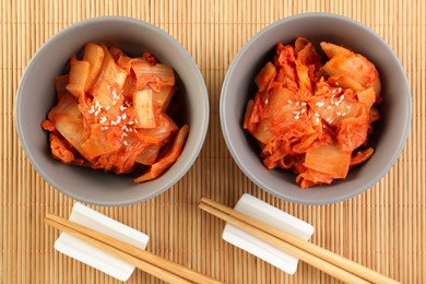 Photo of Bowls of spicy cabbage kimchi and chopsticks on bamboo mat, flat lay