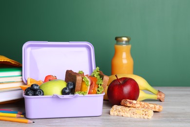 Lunch box with healthy food and different stationery on light wooden table near green chalkboard