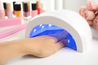 Woman using ultraviolet lamp to dry gel nail polish at white table, closeup