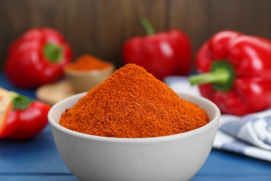 Photo of Bowl with aromatic paprika powder and fresh bell peppers on blue table, closeup