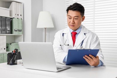 Photo of Doctor with laptop and clipboard consulting patient at white desk in clinic. Online medicine