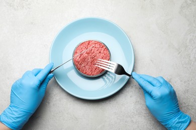 Scientist holding fork and knife over plate with minced cultured meat at light grey lab table, top view